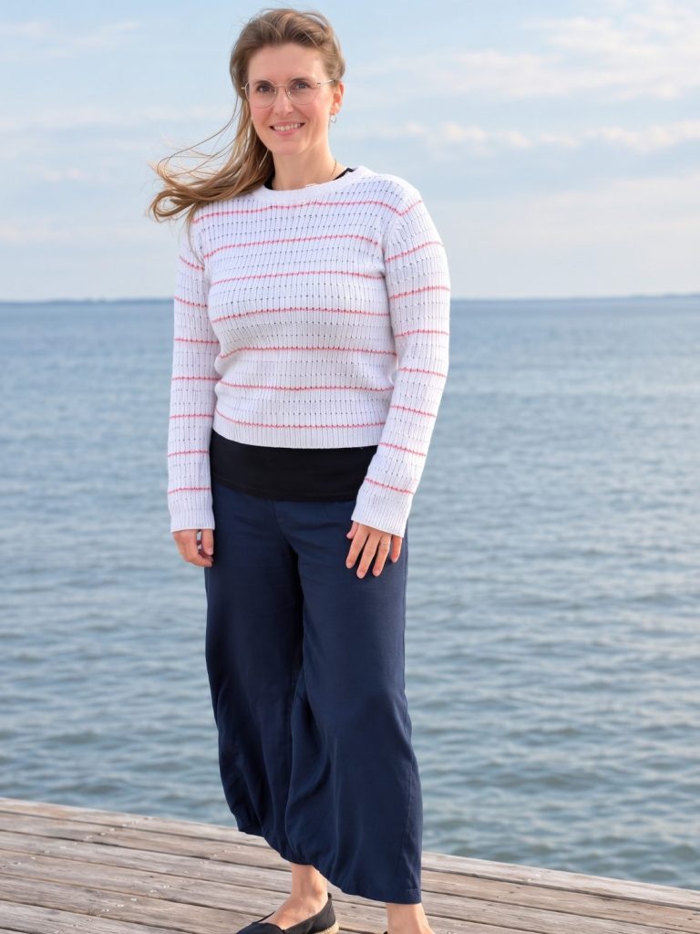 Eine Frau mit blonden Haaren sitzt am Wasser, Enten schwimmen im Hintergrund.